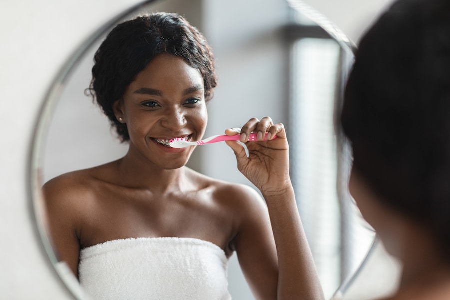 young woman smiling while brushing her teeth with braces and looking in the mirror